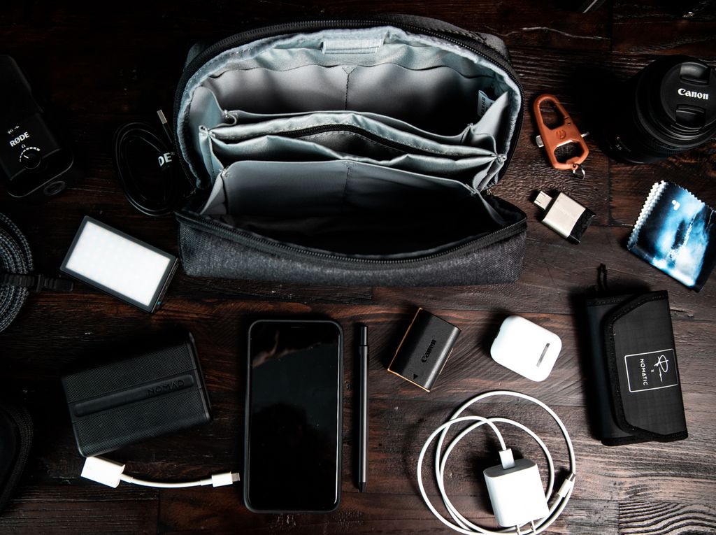 Top view of arranged tools including smartphone camera lens charging flash drives wireless headphones in case placed near bag