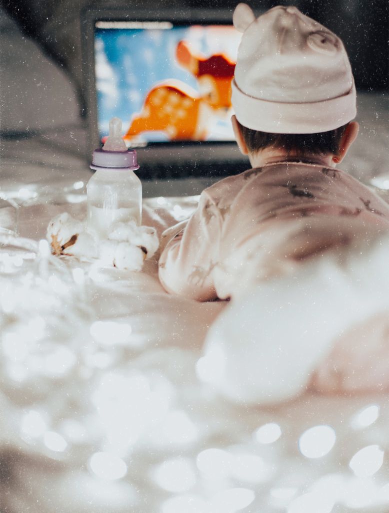 A baby in pajamas watches an animated movie on a laptop, surrounded by soft lights and a bottle At Christmas time 