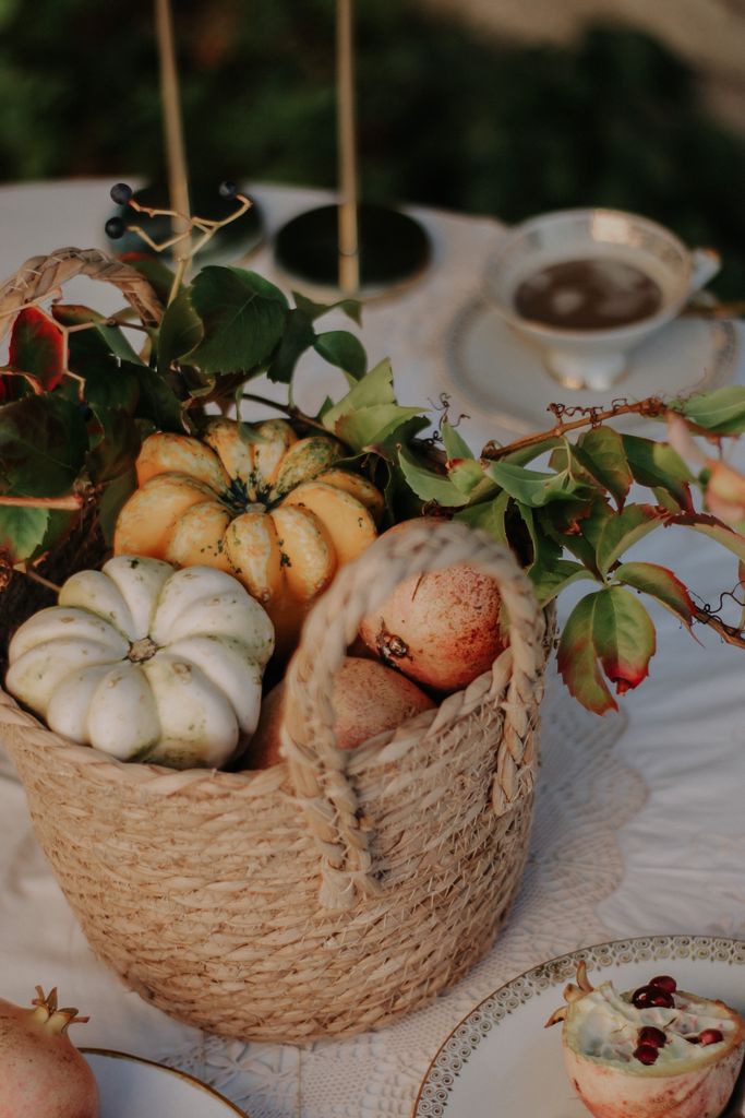 A woven basket filled with pumpkins and fruits, set on a table with elegant tableware.