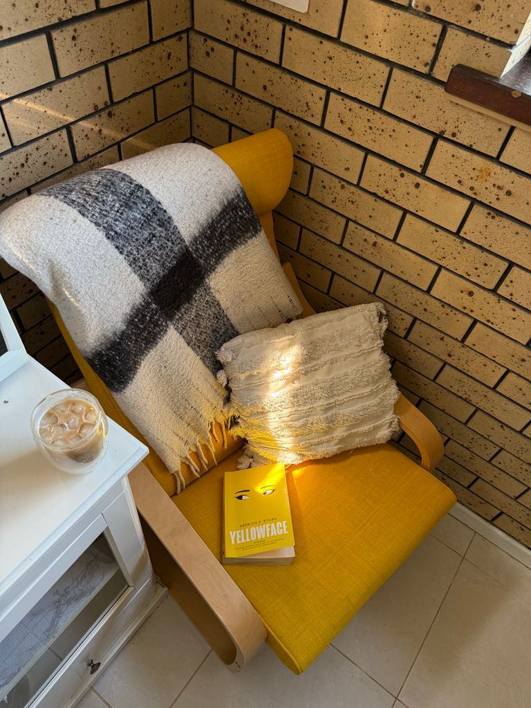 Bright corner armchair with blanket, pillow, book 'Yellowface', and iced coffee.