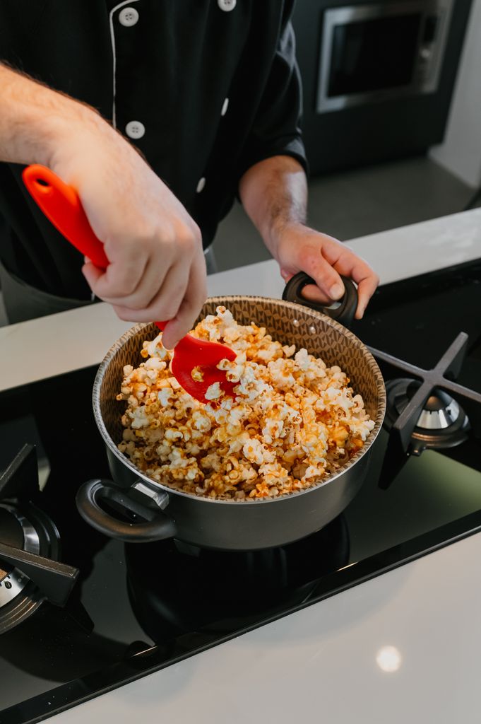 Chef prepares flavored popcorn in a pot on a sleek stovetop, adding seasoning with a red spatula for a Christmas Movie Night in