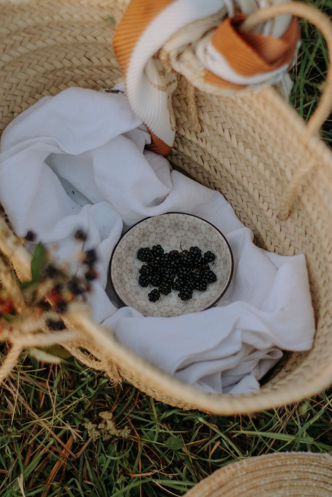 Close-up of blackberries in a wicker basket with white fabric in a grassy picnic setting.