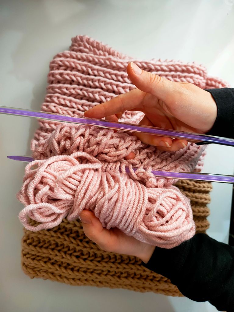 Close-up of hands knitting with pink yarn and needles, showcasing craft and cozy textile work.