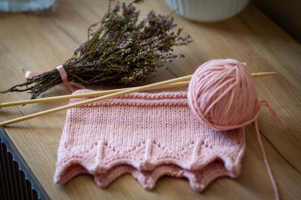 Close-up of pink knitted piece with yarn and needles on a wooden table, accompanied by flowers.
