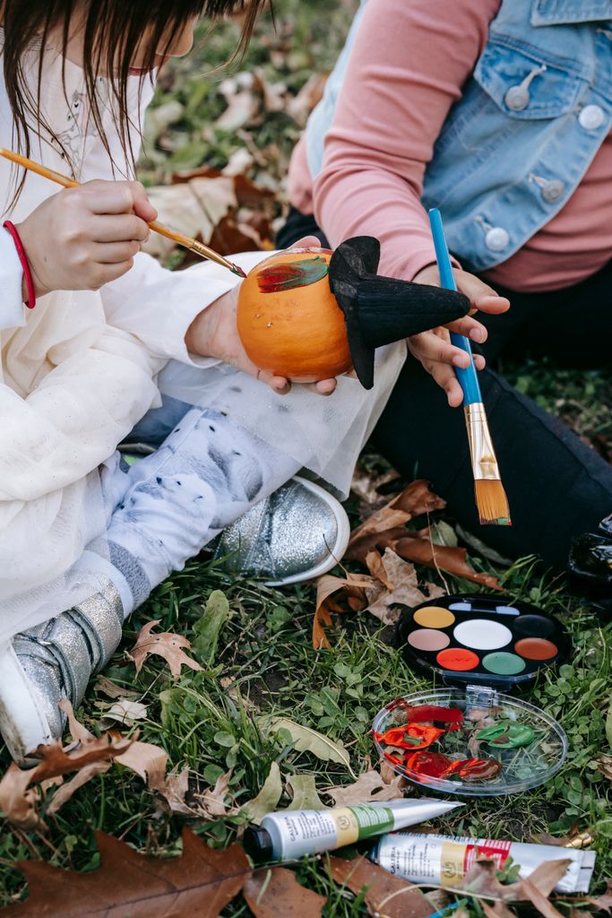 From above unrecognizable girls wearing casual clothes painting Halloween pumpkin while sitting on grass together in October park