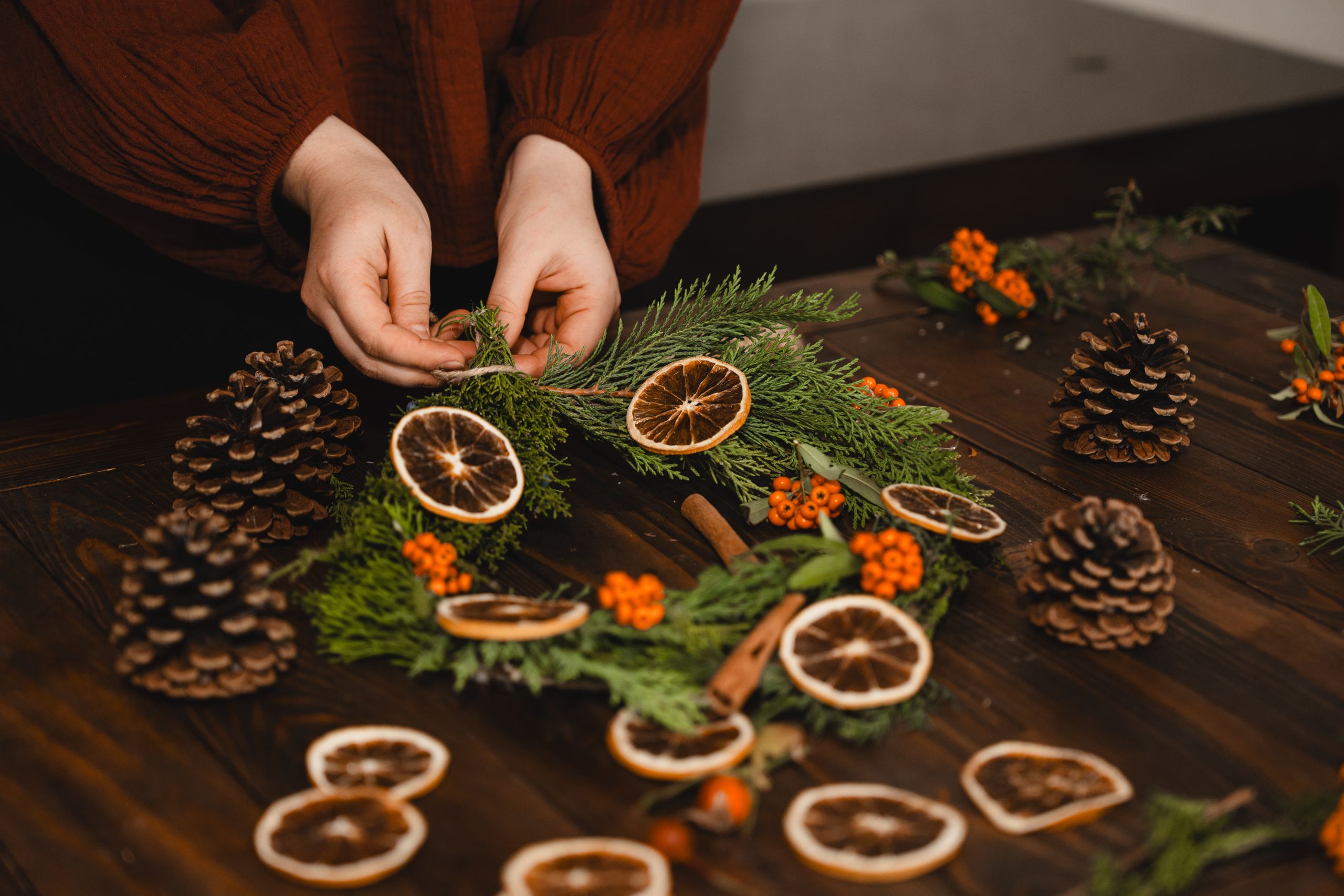 Hands crafting a holiday wreath with dried orange slices, pinecones, and greenery.