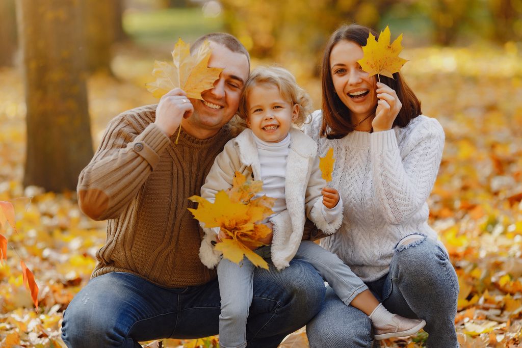Smiling adult parents with adorable little daughter in warm clothes sitting together on ground in autumn park and hiding eyes behind yellow maple leaves and looking at camera