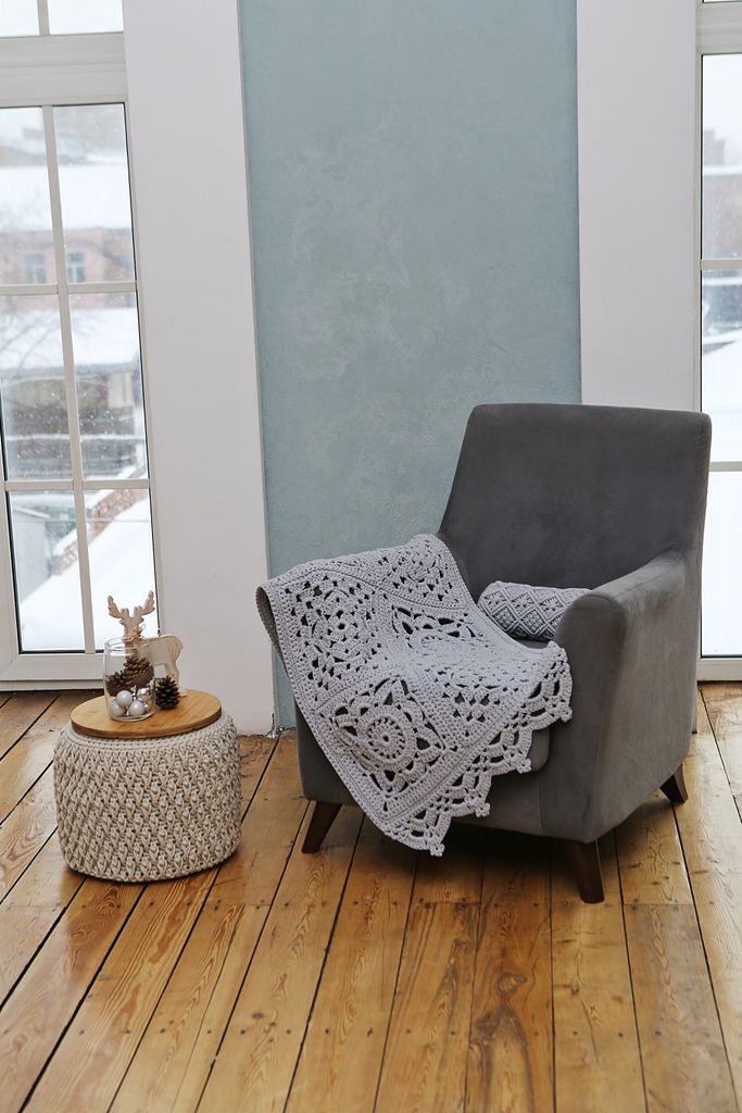 Stylish indoor setup with armchair, crochet blanket, and small table beside snowy window.