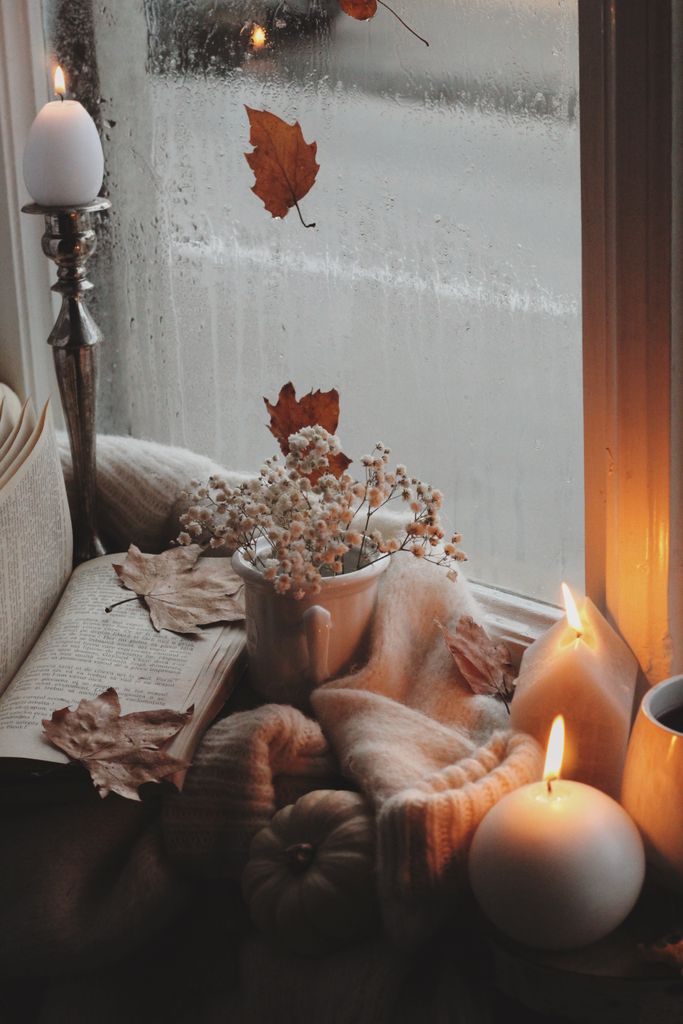 Warm and inviting autumn setup with candles, leaves, and a book by a rainy windowsill.