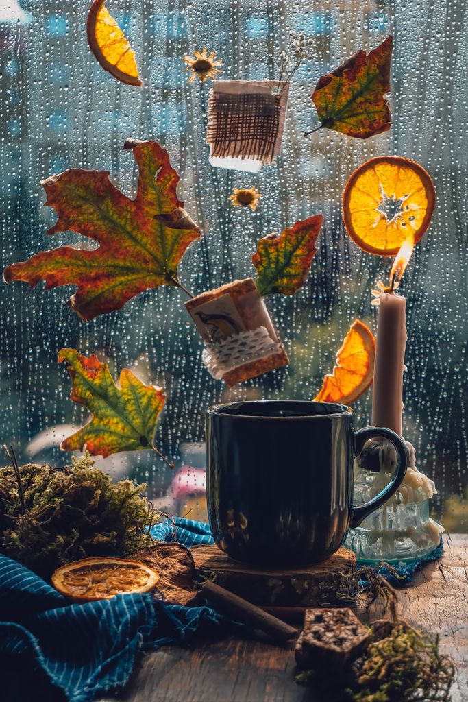 Warm autumn setup with a mug and candle by a rainy window adorned with leaves and orange slices.