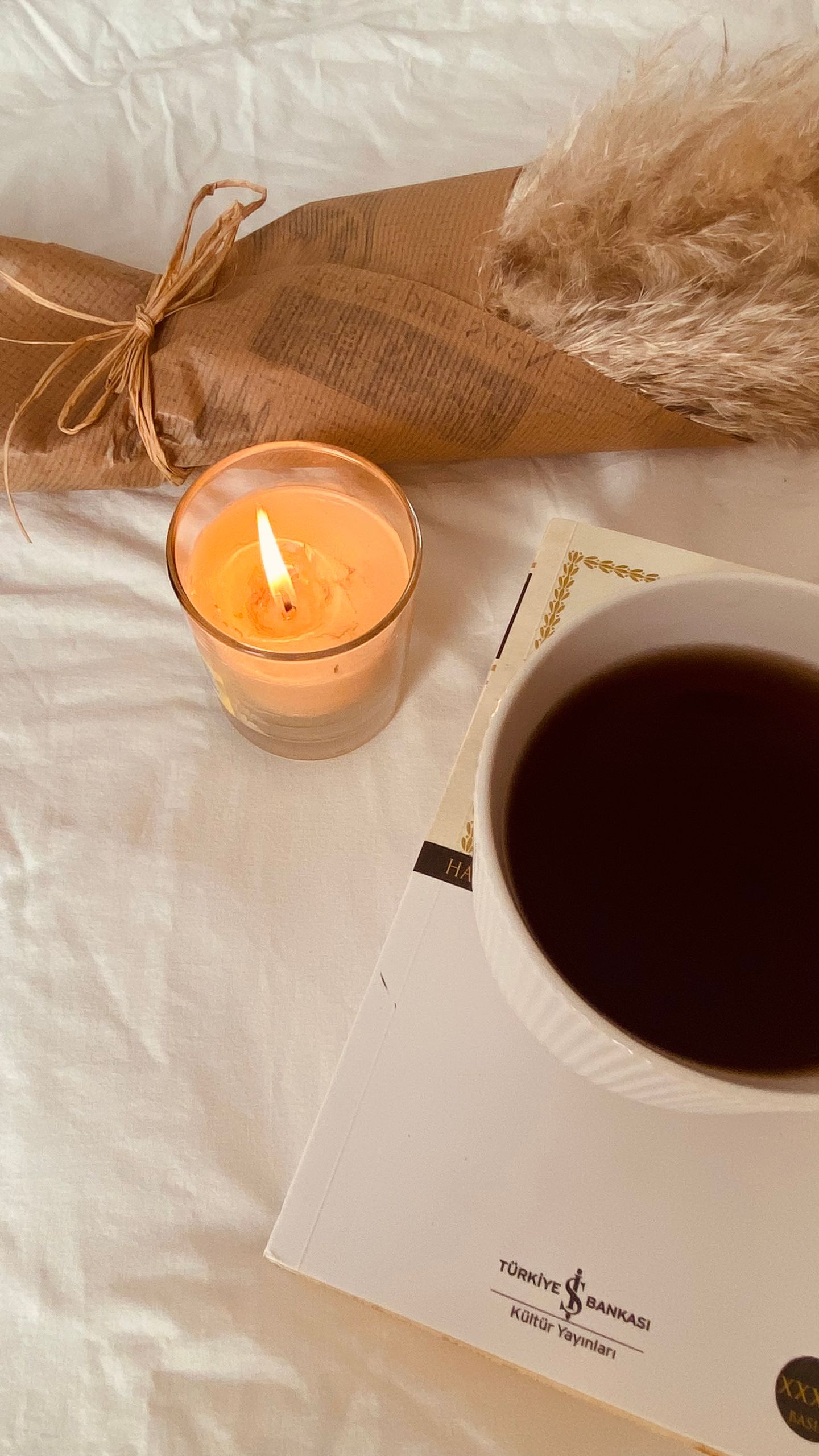 Warm flat lay of coffee, candle, and book on white fabric.