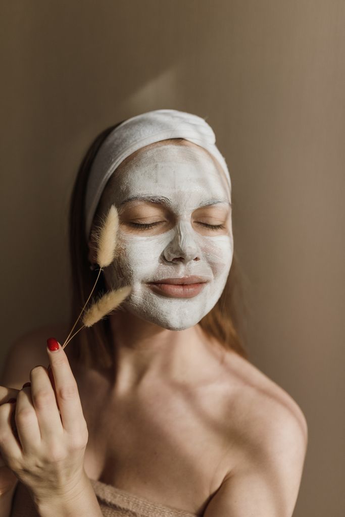 Woman with facial mask and headband enjoying a relaxing skincare treatment indoors.