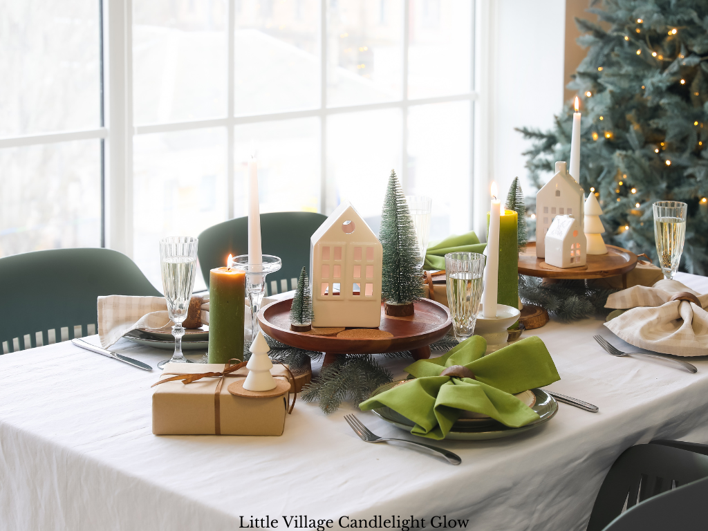 Candlelit Christmas table centerpiece with white porcelain village houses on wooden pedestal stands, bottle brush trees, taper candles and green pillar candles