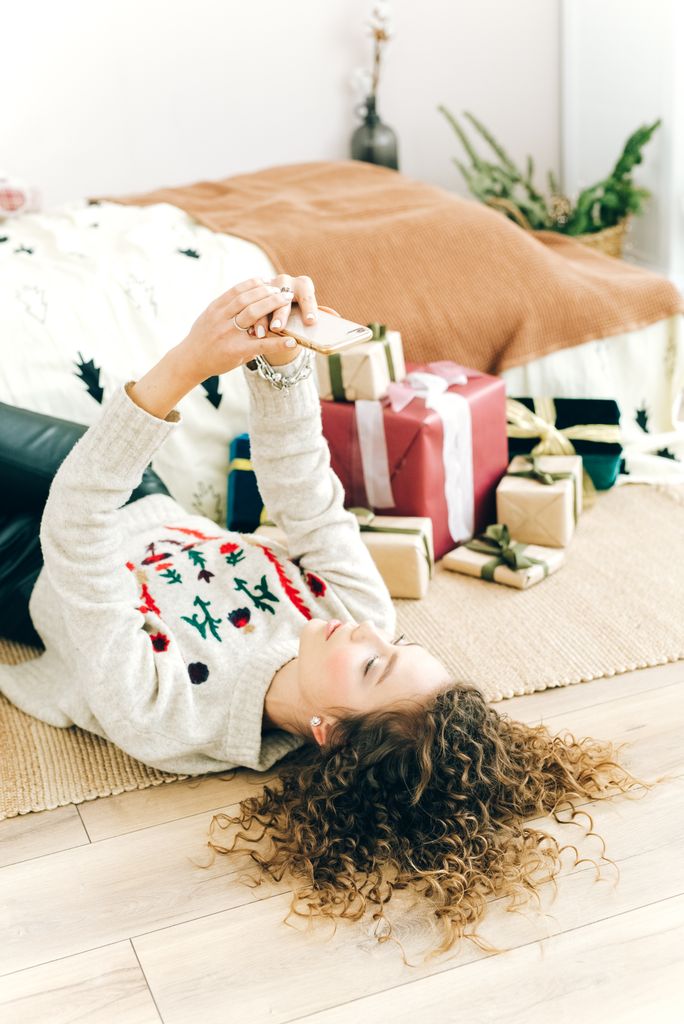A woman with curly hair lying down indoors, taking a selfie beside holiday gifts.