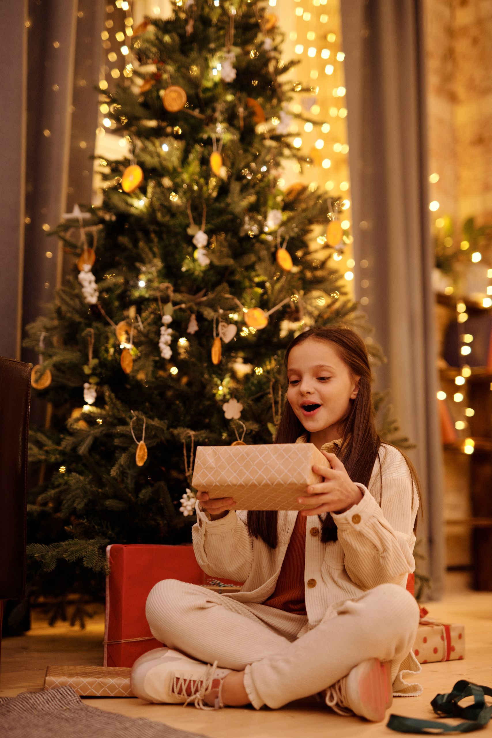 Happy child with present box sitting with crossed legs on floor near shiny decorative tree during New Yer holiday